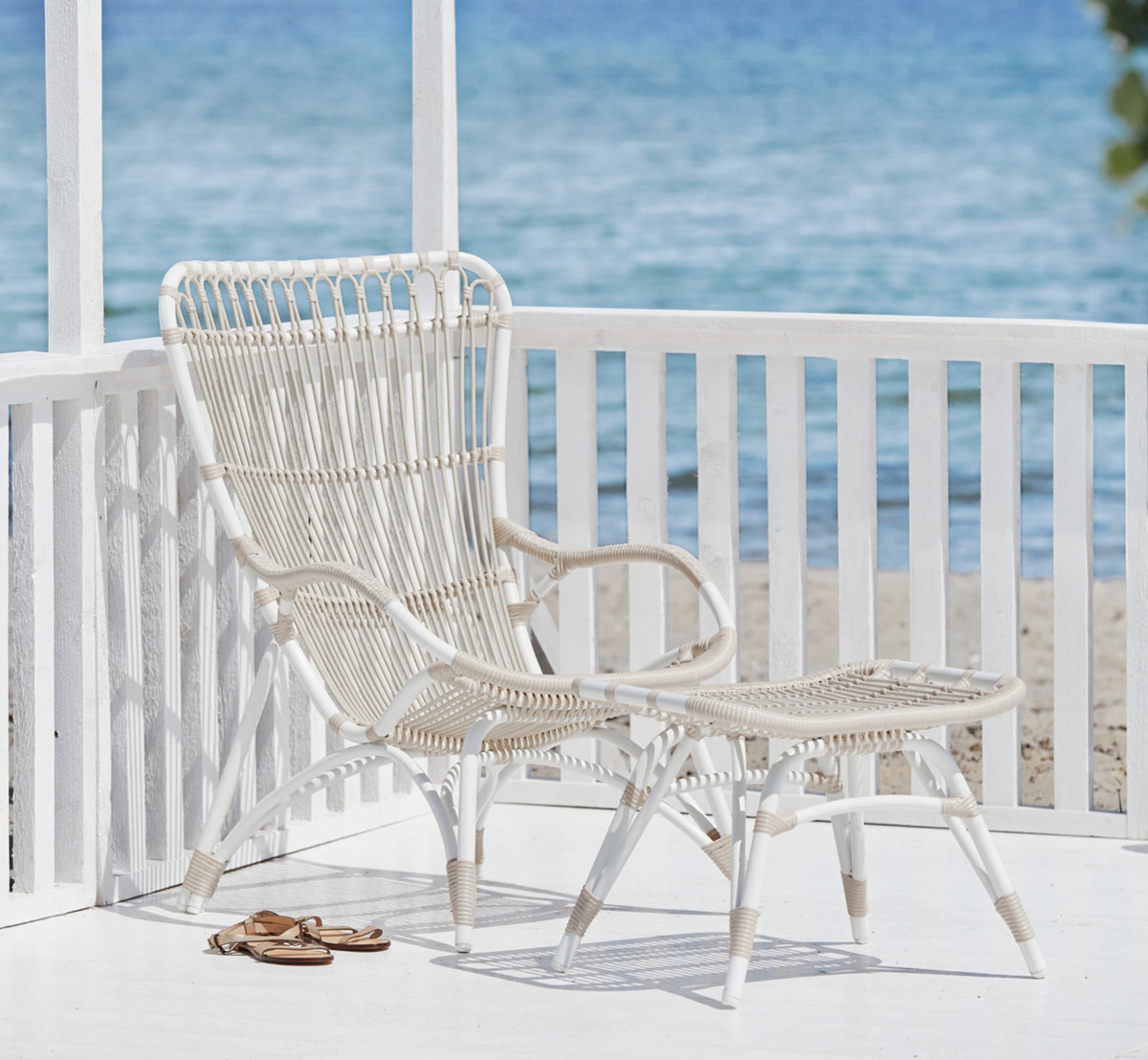 Dies ist ein Lifestyle-Bild von Monet Exterior Footstool i dove white aluminium artfibre. Mit Lounge-Sessel auf einer Veranda am Meer.