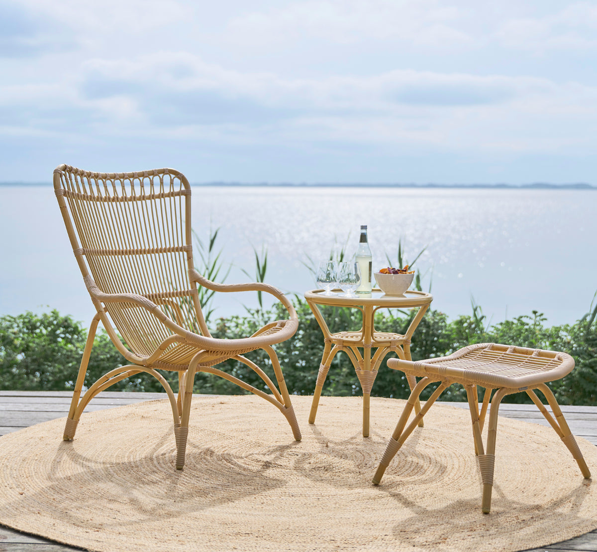 Dies ist ein Lifestyle-Bild von Monet Exterior Footstool i natural aluminium artfibre. Mit Lounge-Sessel auf einem Deck am Meer.
