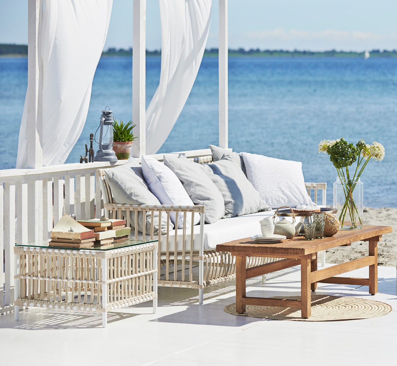 Dies ist ein Lifestyle-Bild von Caroline Außen-Sofa in dove White Aluminium ArtFibre, auf einem Holzdeck am Wasser mit Küstengrün und Meerblick.