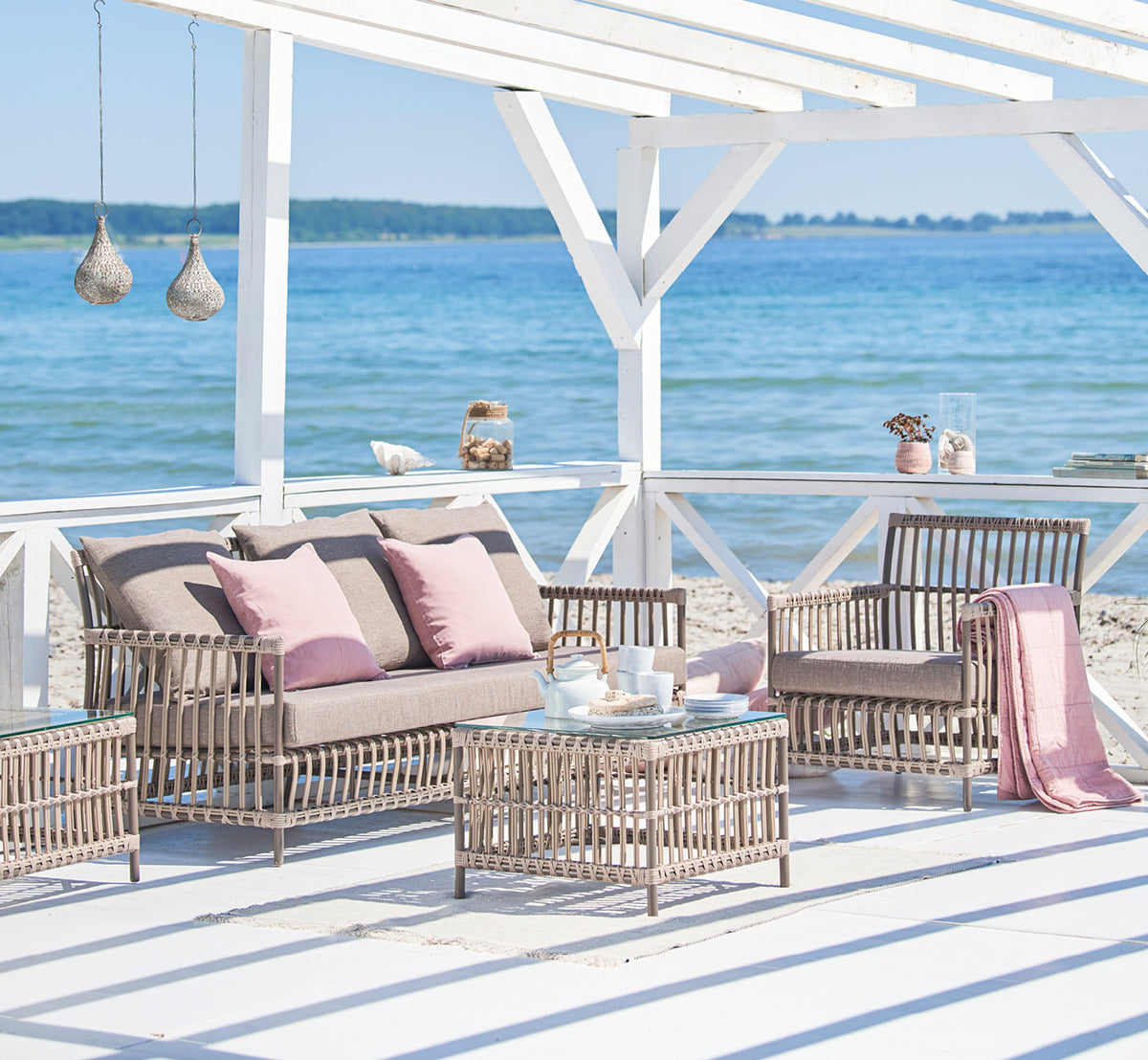 Dies ist ein Lifestyle-Bild von Caroline Außen-Sofa in moccaccino Aluminium ArtFibre, auf einem Holzdeck am Wasser mit Küstengrün und Meerblick.