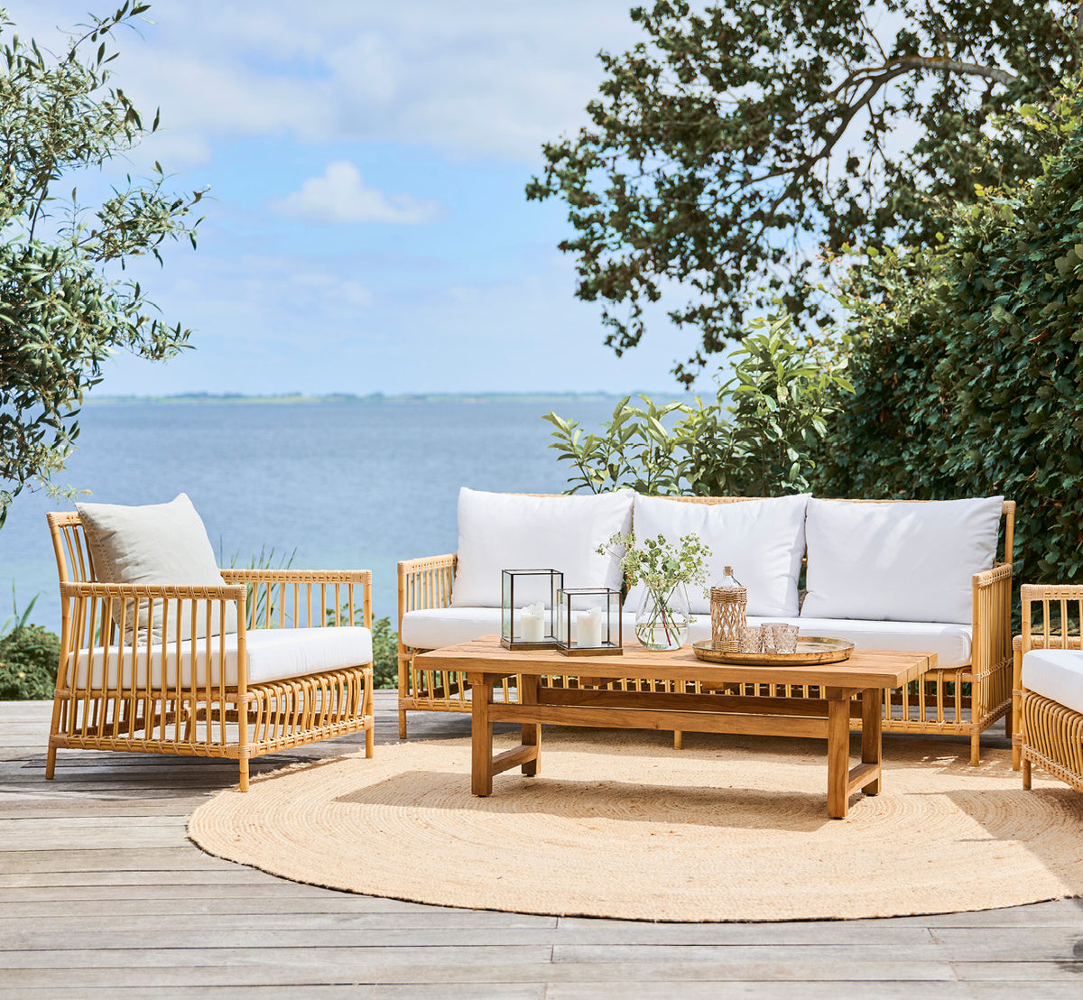 Dies ist ein Lifestyle-Bild von Caroline Außen-Sofa in natural Aluminium ArtFibre, auf einem Holzdeck am Wasser mit Küstengrün und Meerblick.