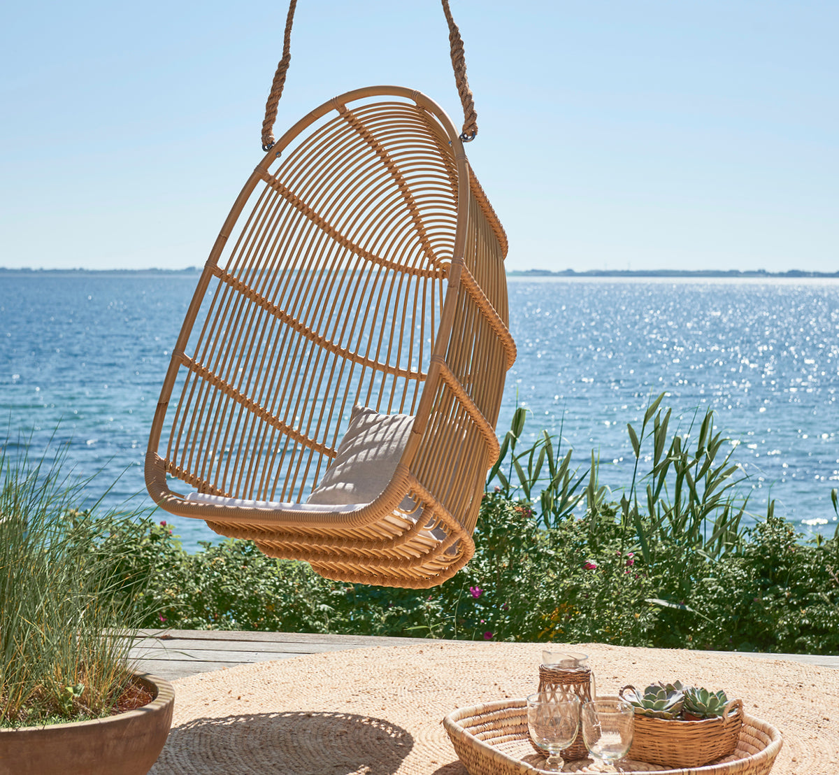 Dies ist ein Lifestyle-Bild von Renoir Außen-Hängesessel in natural aluminium artfibre. Auf einer Strandterrasse mit Tablett und Sukkulenten darunter hängend.