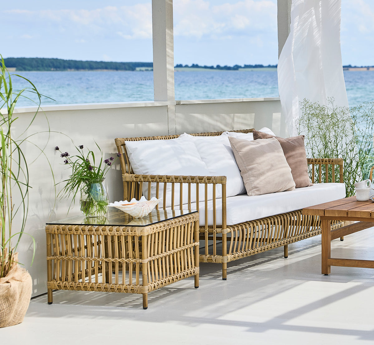 Dies ist ein Lifestyle-Bild von Caroline Außentisch in antique Aluminium ArtFibre, auf einem Holzdeck am Wasser mit Küstengrün und Meerblick.