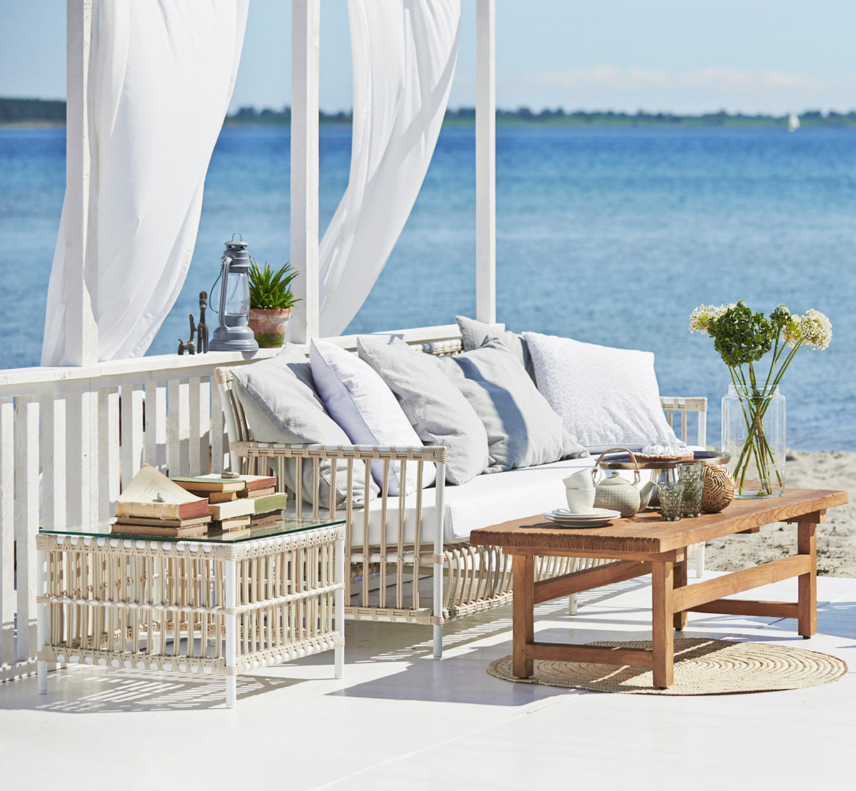 Dies ist ein Lifestyle-Bild von Caroline Außentisch in dove White Aluminium ArtFibre, auf einem Holzdeck am Wasser mit Küstengrün und Meerblick.