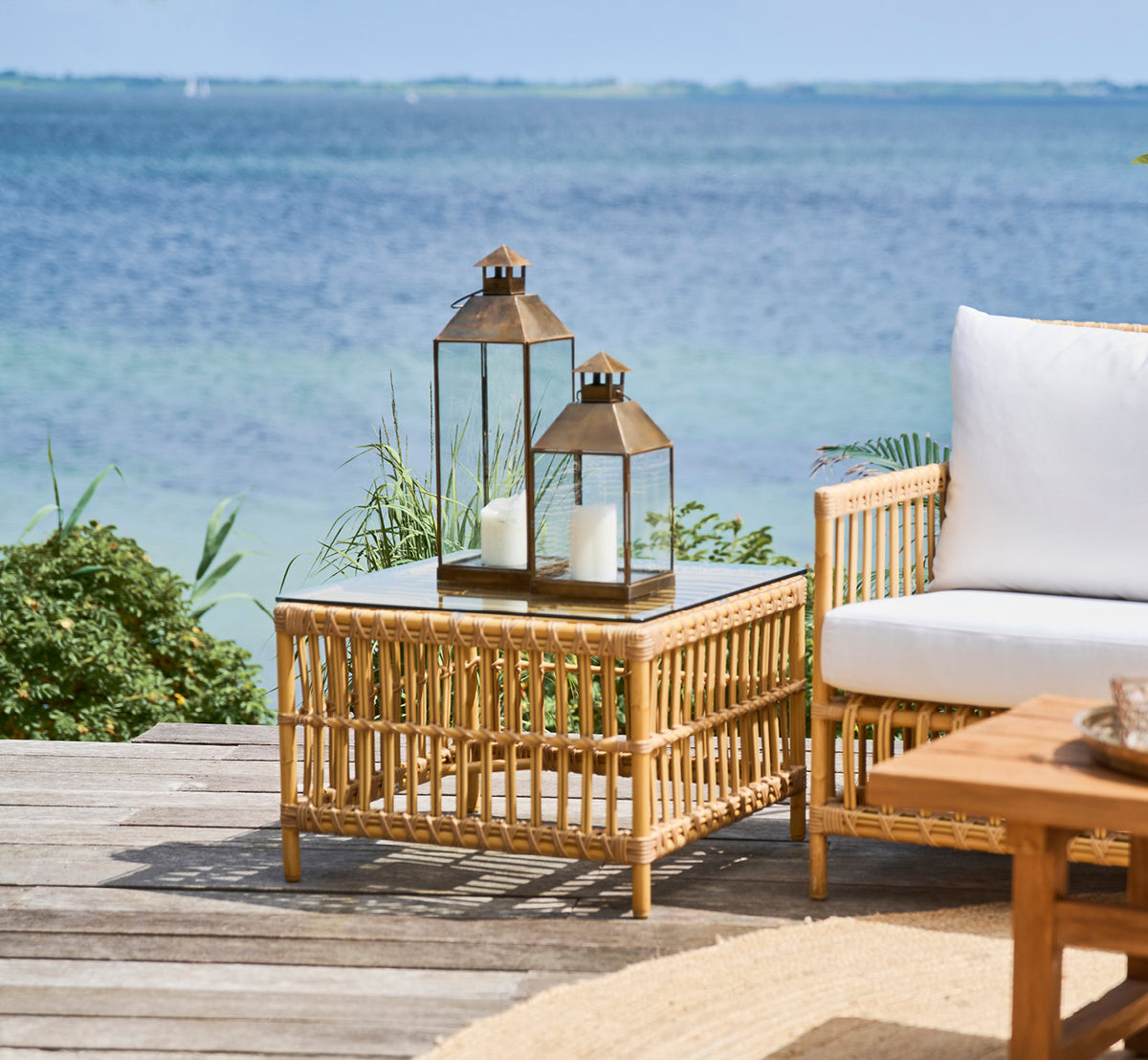 Dies ist ein Lifestyle-Bild von Caroline Außentisch in natural Aluminium ArtFibre, auf einem Holzdeck am Wasser mit Küstengrün und Meerblick.