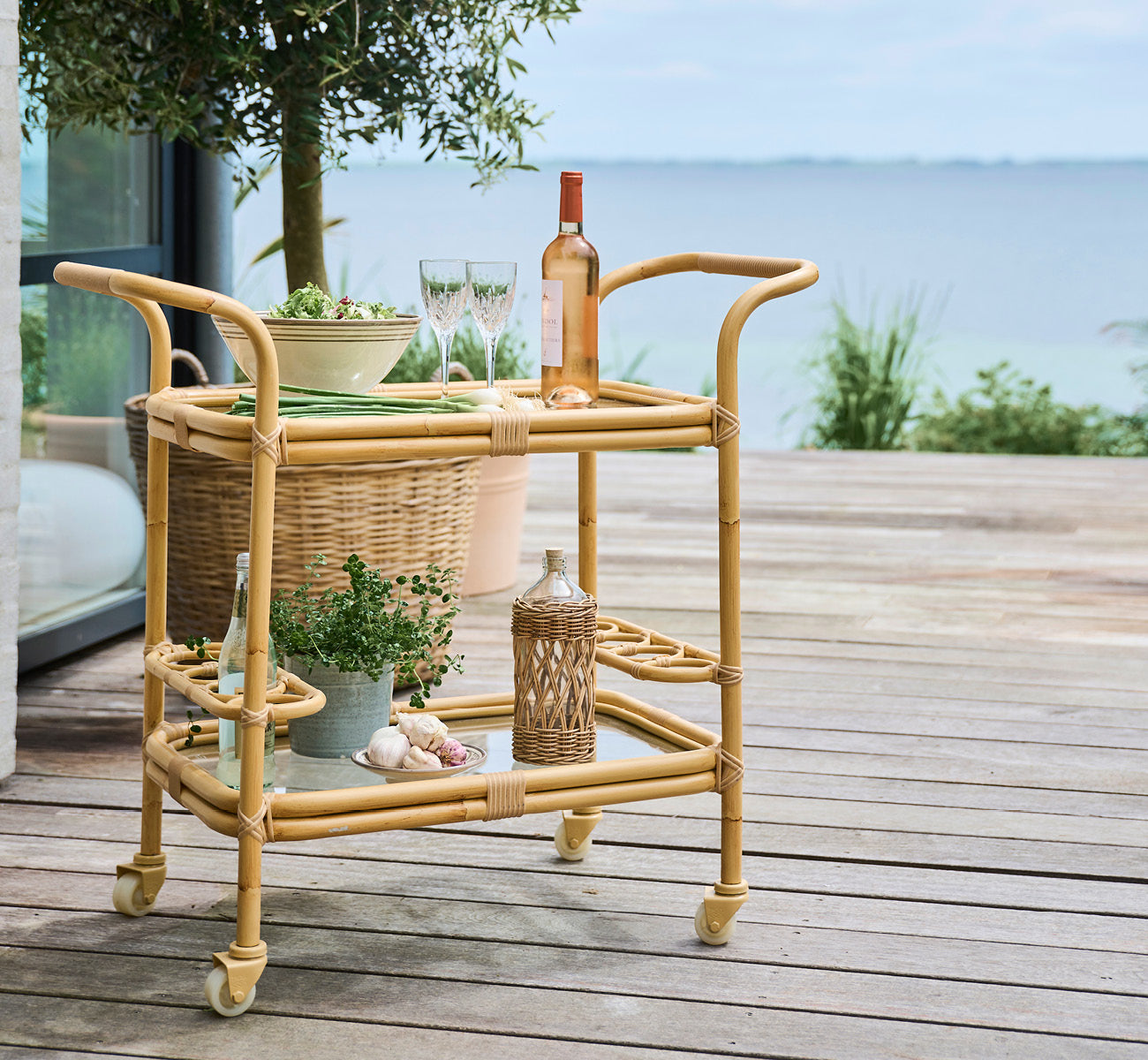 Dies ist ein Lifestyle-Bild von Carlo Außen-Servierwagen in natural Aluminium ArtFibre, auf einem Holzdeck am Wasser mit Küstengrün und Meerblick.