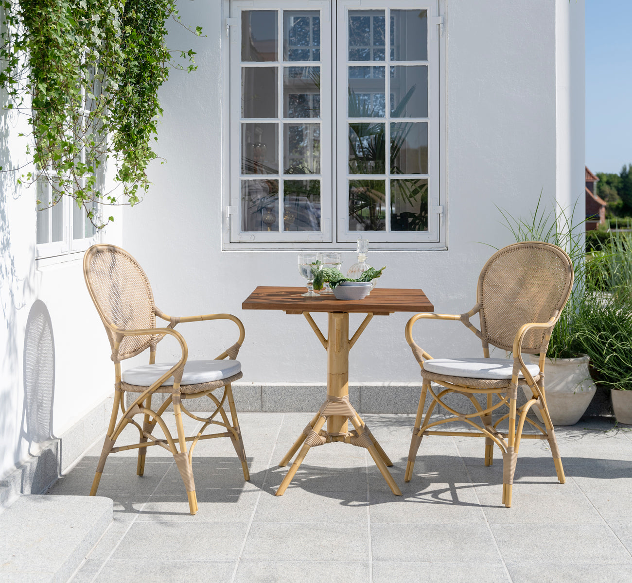 Dies ist ein Lifestyle-Bild von Nicole Cafe Table i natural aluminium artfibre. Auf einer Steinterrasse mit Ofelia-Stühlen und Grünpflanzen.