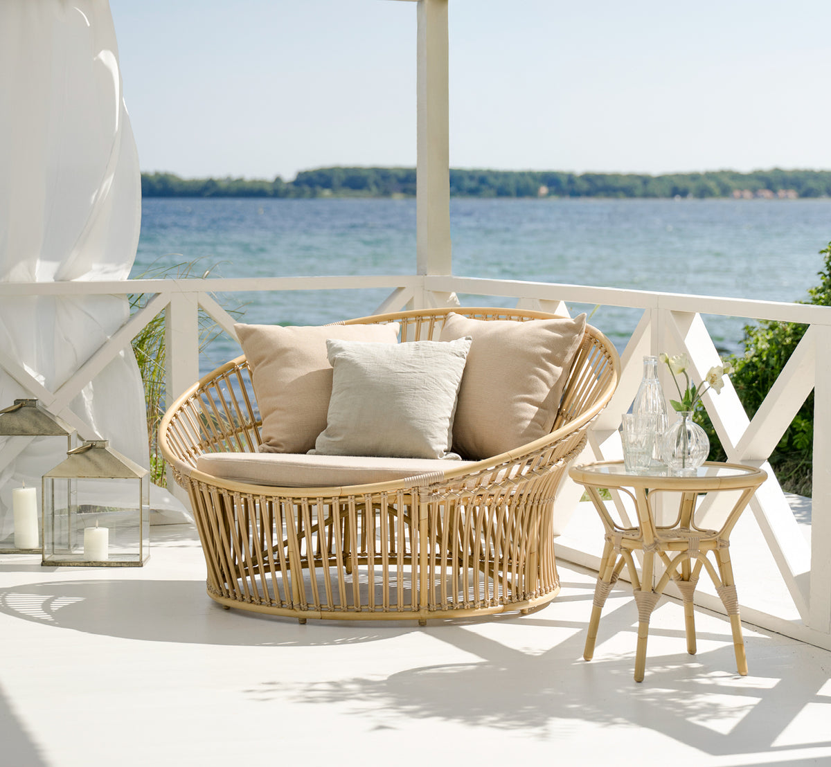 Dies ist ein Lifestyle-Bild von Olympia Nest Exterior i natural aluminium artfibre. In einem Pavillon am Meer mit Leinenkissen.