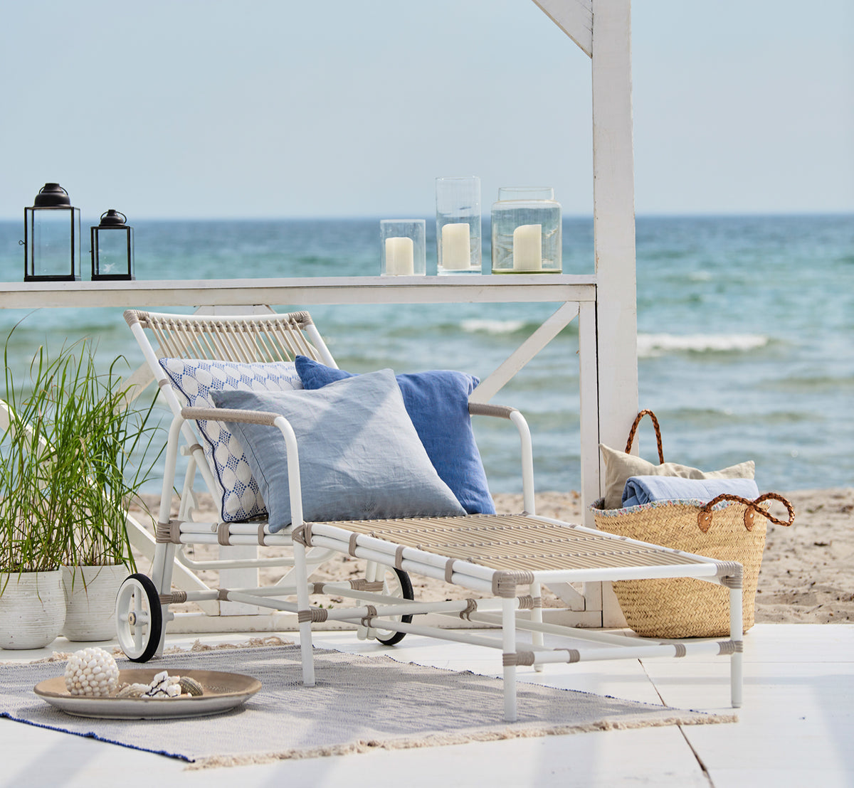 Dies ist ein Lifestyle-Bild von Josephine Sonnenliege in dove White aluminium artfibre, auf einem weißen Holzdeck am Strand unter einer weißen.