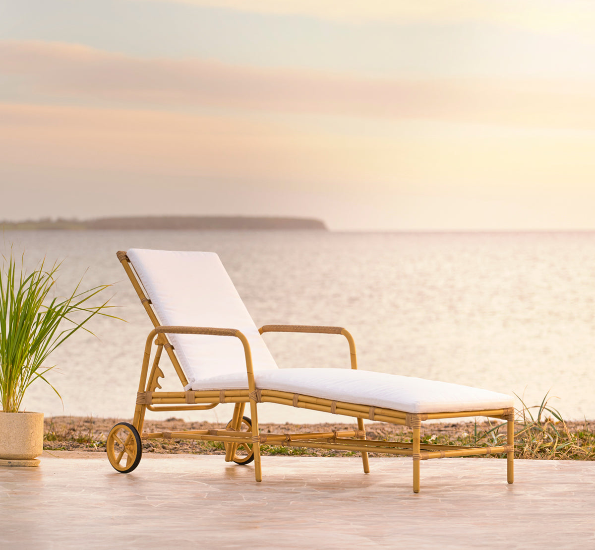 Dies ist ein Lifestyle-Bild von Josephine Sonnenliege in natural aluminium artfibre, auf einer Steintedrasse an der Küste bei Sonnenuntergang mit.