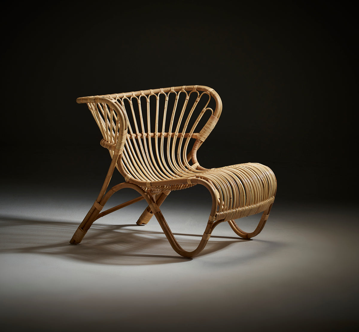 Dies ist ein Lifestyle-Bild von Fox Lounge-Sessel in polished Natural Rattan, in einem warmen Interieur mit ockerfarbener Wandvertäfelung und dunklem.