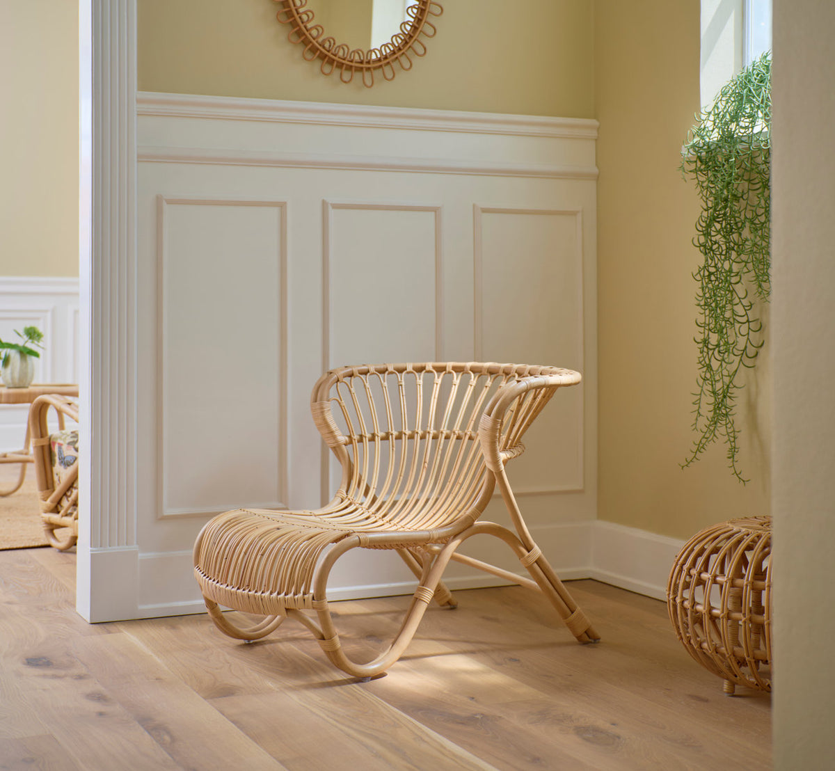 Dies ist ein Lifestyle-Bild von Fox Lounge-Sessel in polished natural rattan. Vor weißen Paneelen mit einem Rattanspiegel.