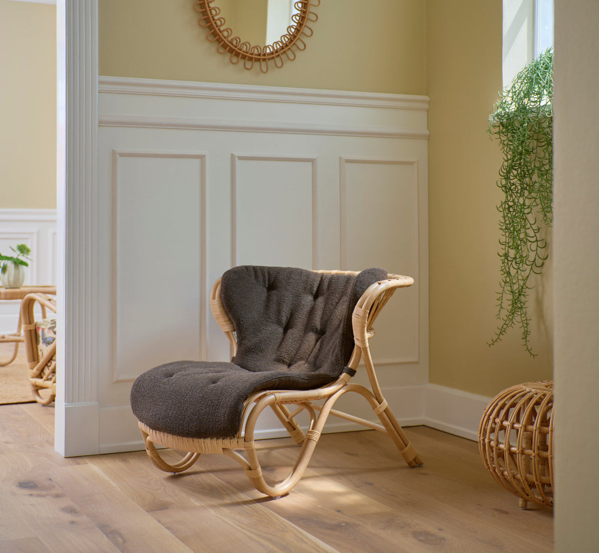 Dies ist ein Lifestyle-Bild von Fox Lounge-Sessel in polished natural rattan. Mit grauem Kissen vor weißen Paneelen.