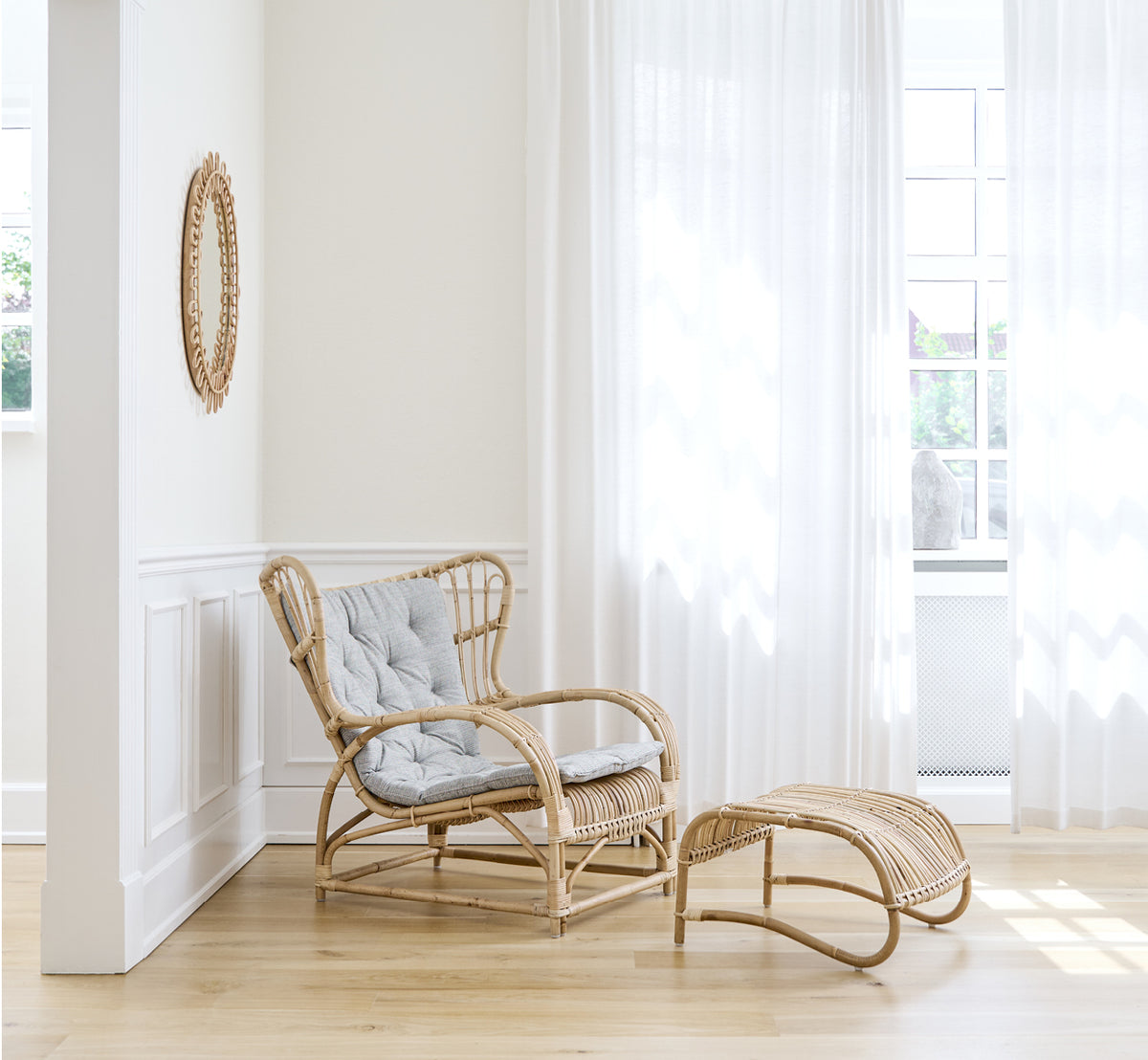 Dies ist ein Lifestyle-Bild von Teddy Lounge-Sessel in polished natural rattan. Auf einem Fischgrätboden an einem Vorhangfenster mit kleinem Spiegel platziert.