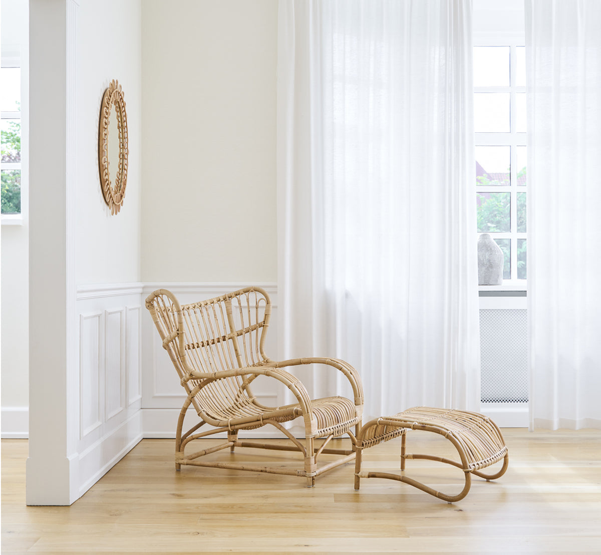 Dies ist ein Lifestyle-Bild von Teddy Fußhocker in polished natural rattan. Mit dem passenden Loungesessel an einem hellen Vorhangfenster kombiniert.