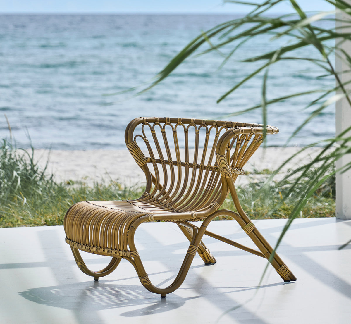 Dies ist ein Lifestyle-Bild von Fox Außen-Lounge-Sessel in antique Aluminium ArtFibre, auf einem Holzdeck am Wasser mit Küstenschilf und Meerblick.