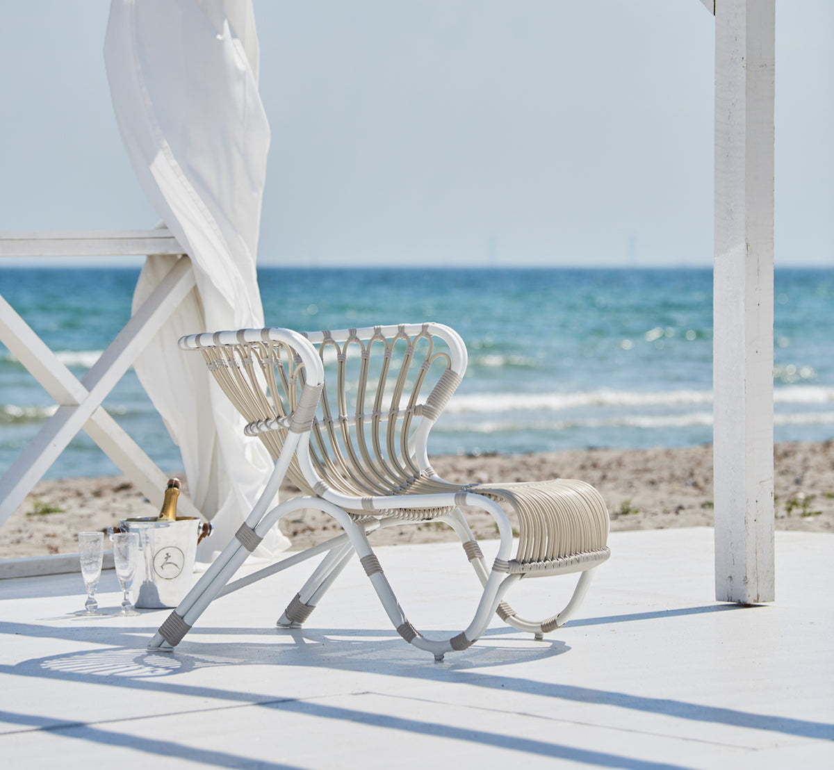 Dies ist ein Lifestyle-Bild von Fox Außen-Lounge-Sessel in dove white Aluminium ArtFibre, auf einem Holzdeck am Wasser mit Küstenschilf und Meerblick.