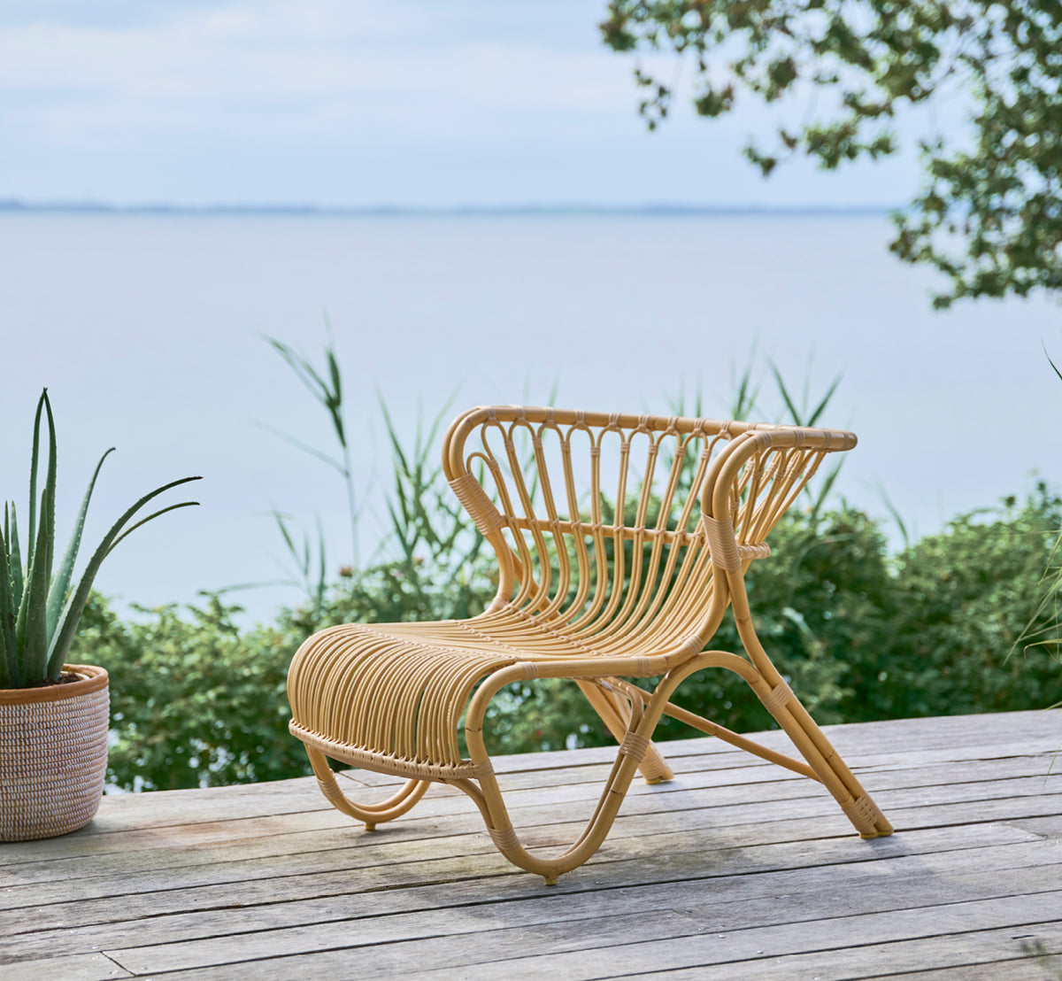Dies ist ein Lifestyle-Bild von Fox Außen-Lounge-Sessel in natural Aluminium ArtFibre, auf einem Holzdeck am Wasser mit Küstenschilf und Meerblick.