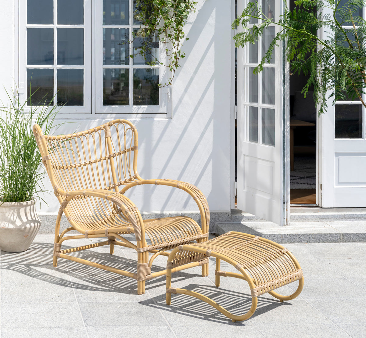 Dies ist ein Lifestyle-Bild von Teddy Außen-Lounge-Sessel in natural aluminium artfibre. Auf einer sonnigen gepflasterten Terrasse an einem weiß gerahmten Türrahmen platziert.