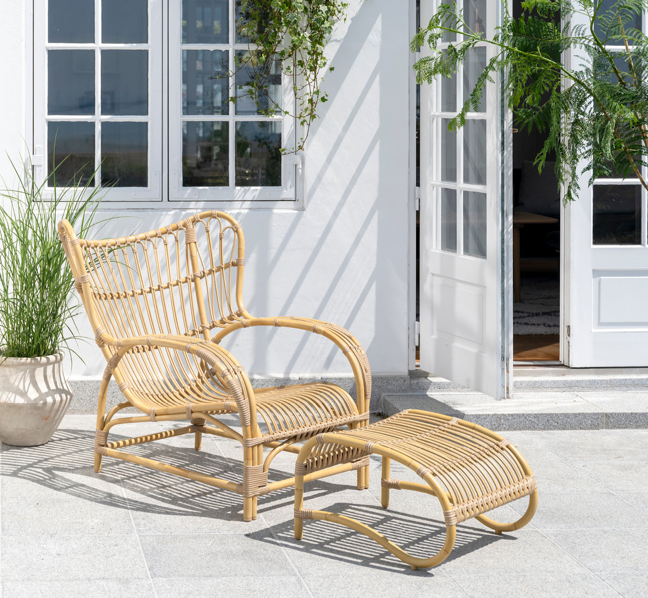 Dies ist ein Lifestyle-Bild von Teddy Außen-Lounge-Sessel in natural aluminium artfibre. Auf einer sonnigen gepflasterten Terrasse an einem weiß gerahmten Türrahmen platziert.