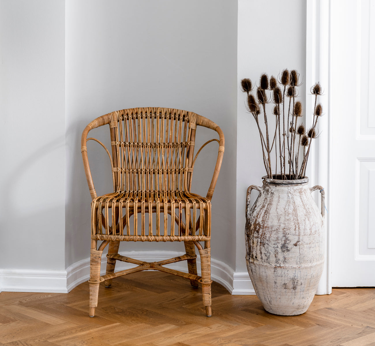 Dies ist ein Lifestyle-Bild von Robert Esszimmerstuhl in polished antique rattan. Neben einer hohen Terrakotta-Urne auf einem Fischgrätparkettboden platziert.