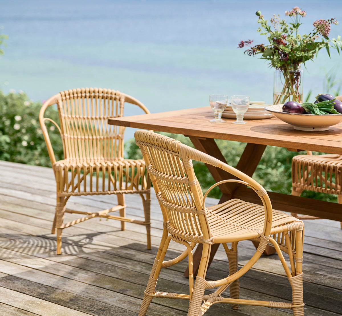 Dies ist ein Lifestyle-Bild von Robert Außen-Esszimmerstuhl in natural aluminium artfibre. Um einen Teaktisch auf einer Holzterrasse mit Meerblick angeordnet.