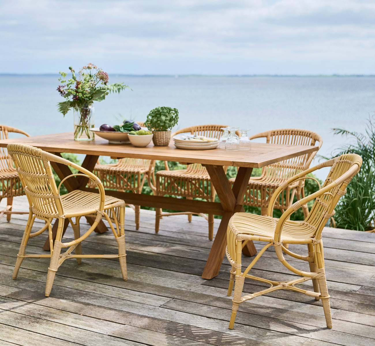Dies ist ein Lifestyle-Bild von Robert Außen-Esszimmerstuhl in natural aluminium artfibre. Um einen Teaktisch auf einer Holzterrasse mit Meerblick angeordnet.