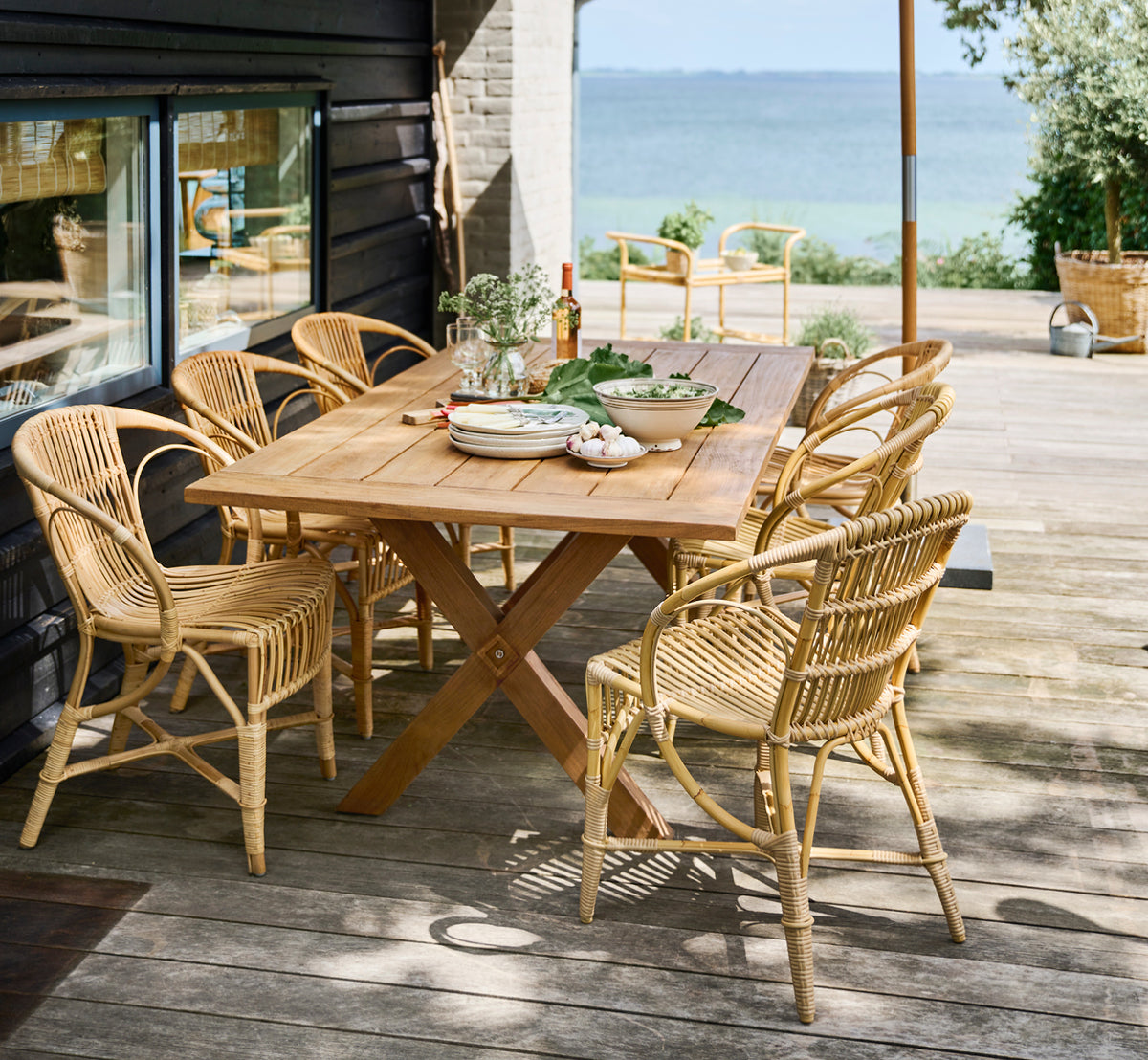 Dies ist ein Lifestyle-Bild von Robert Außen-Esszimmerstuhl in natural aluminium artfibre. Um einen Teaktisch auf einer Holzterrasse mit Meerblick angeordnet.