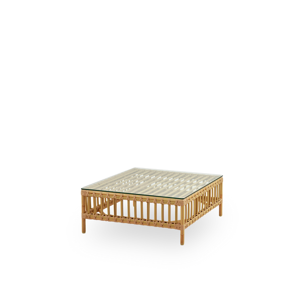 Seitenansicht des Maggie Outdoor-Sofas Pouf-Moduls in Natural-Farbe, präsentiert mit Glasplatte zur Nutzung als Beistelltisch. Aluminiumrahmen im handbemalten Rattan-Look mit ArtFibre-Umschlingungen.
